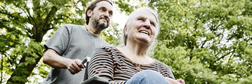 Eine ältere Frau sitzt im Rollstuhl und wird von einem jungen Mann angeschoben
