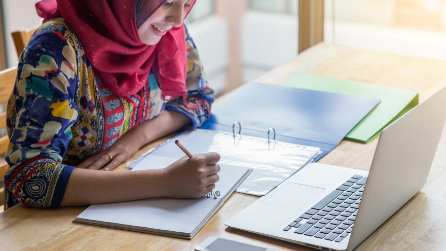 Junge Frau mit pinkem Kopftuch sitzt mit Unterlagen vor ihrem Laptop