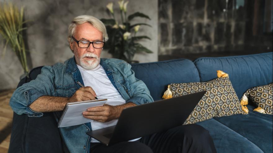 älterer Mann hat Laptop auf dem Schoß und macht Notizen