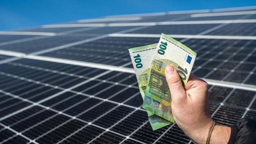 Eine männliche Hand hält zwei Hundert-Euro-Scheine in der Hand vor einer Photovoltaikanlage