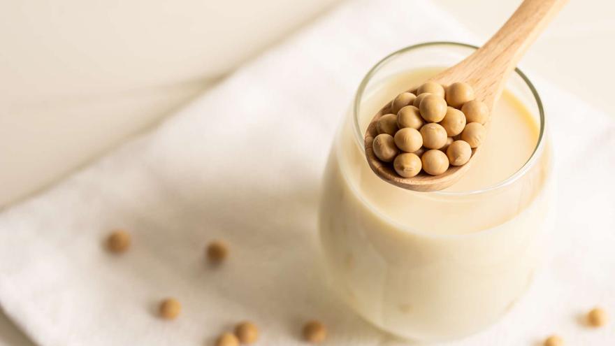 Ein Glas cremige Sojamilch steht im Zentrum dieser hellen, minimalistischen Komposition auf einem weißen Stoffuntergrund, während ein darüber balancierter Holzlöffel eine Häufung runder, gelblicher Sojabohnen präsentiert. Die sanfte Beleuchtung in warmen Beigetönen betont die natürliche Textur der Zutaten, die durch weitere dekorativ verstreute Bohnen im weichgezeichneten Hintergrund ergänzt werden.