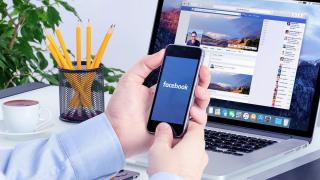 Mann am Schreibtisch hält Smartphone mit dem Schriftzug facebook auf dem Display in seinen Händen, dahinter ein aufgeklappter Laptop mit geöffneter Facebook-Seite