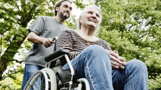 Eine ältere Frau sitzt im Rollstuhl und wird von einem jungen Mann angeschoben